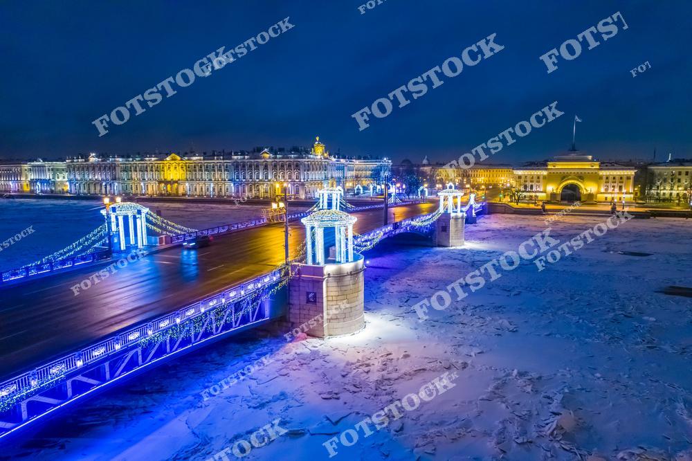 Панорамная фотография зимнего Санкт-Петербурга, снятая с высоты птичьего полета в вечернее время. На снимке запечатлен Дворцовый мост, украшенный праздничной подсветкой, замерзшая Нева и здание Эрмитажа в огнях ночного города. Атмосфера зимнего Петербурга передается холодными оттенками льда, яркими огнями архитектурных ансамблей и отражением света на снежной поверхности. Вид с дрона позволяет увидеть масштаб исторического центра, мосты и знаменитые достопримечательности Северной столицы России. Фотография идеально подходит для туристических публикаций, фотостоков, новогодней рекламы, оформления интерьеров и материалов о зимнем Санкт-Петербурге.